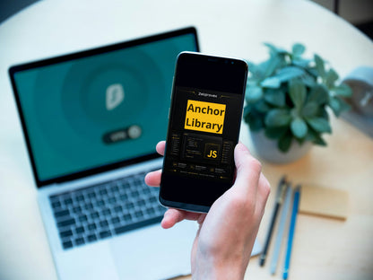 A hand holding a smartphone displaying the 'Anchor Library' JS library, with an open laptop in the background on a light-colored desk next to a small potted plant.