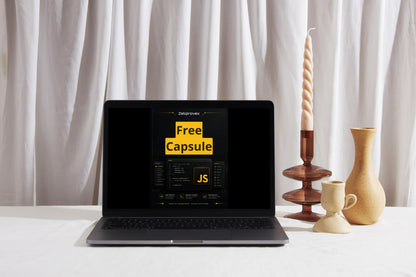 A black laptop sits open on a white surface in front of draped white curtains, displaying a website template titled 'Free Capsule' with yellow and white text on the screen. To the right of the laptop are decorative objects: a tall twisted candle in a dark wooden holder, a small beige ceramic cup, and a beige ceramic vase.